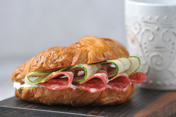 Croissant stuffed with salami, fresh cucumber and cottage cheese. Croissant occupies the entire frame space. In the background is a cup with a drink. Macro shot.