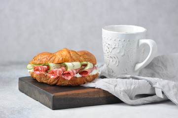Croissant stuffed with salami, fresh cucumber and cottage cheese. Next to the croissant is a cup of...