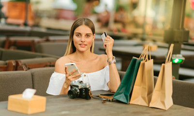 Online shopping. Young shopaholic woman sits at table with shopping bags and holds in her hands smartphone and bank card in outdoor cafe