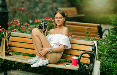 Young fashionable woman in summer clothes holding a retro camera while sitting on bench in the park