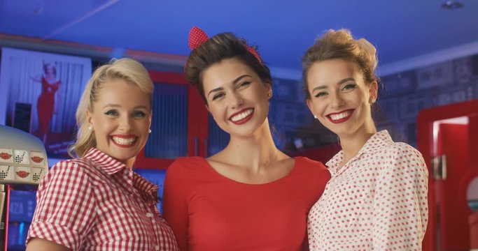 Portrait shot of cute Caucasian beautiful friends, women in retro of 60s style standing in bar and smiling cheerfully. Pretty vintage glamorous girls looking at camera.