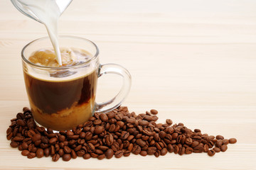 pouring milk into instant coffee in glass mug with roasted coffee beans on wooden background with copy space