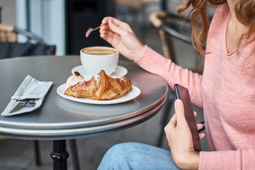 Woman have breakfast in caffe and using smartphone. Girl chatting and using internet with phone during coffee break with a croissant