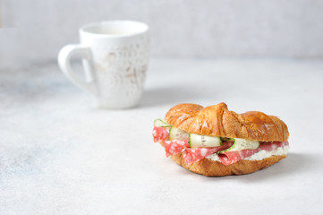 Croissant and a cup of coffee. Croissant filling consists of slices of salami, fresh cucumber and soft curd cheese. Close-up. Light background.