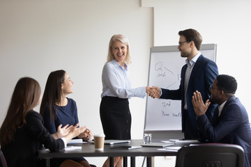 Confident businessman or boss shake hand of excited mature female employee greeting with work success at meeting, smiling male director handshake happy woman worker congratulate with job promotion