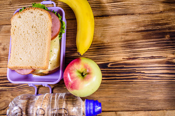 Ripe apple, banana, bottle of water and lunch box with sandwiches on a wooden table. Top view