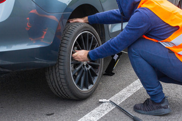 A man in a orange safety vest changes a flat tire on a road. Close-up mans hands to the wheel of a broken car. Replacement of a wheel using skrewdriver.