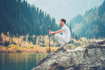 A wise man sits on a stone and holds a sword in his hands. Mountain Lake background