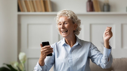 Overjoyed mature 60s woman sit on couch at home triumph reading good news on cell, excited senior...