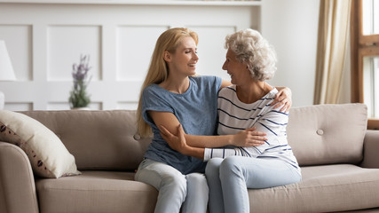 Smiling mature mother and grownup daughter sit relax on couch in living room talking and chatting, happy senior mom and adult girl child rest on sofa at home, enjoy family weekend bonding together