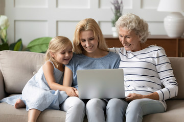 Happy three generations of women sit relax on couch in living room using modern laptop together, smiling little girl with young man and grandmother rest on sofa watching video on computer at home
