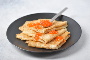 Pancakes with red caviar on a black plate. Next to the pancakes lies a spoon with red caviar. Close-up. Light background