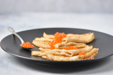 Pancakes on a black plate. On pancakes, red caviar. Next to the pancakes is a spoon with red caviar. Close-up. Light background.