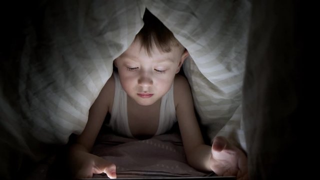 little boy plays on tablet while lying under blanket. watching cartoons hiding