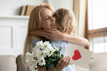 Obraz na płótnie Canvas Cute little loving preschooler daughter make birthday surprise present flowers and handmade postcard to excited young mother, caring small girl child greeting congratulating overjoyed mom or nanny