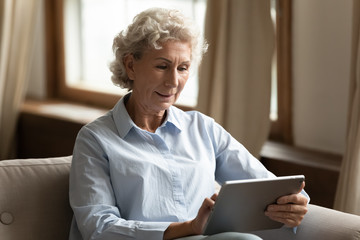 Modern mature 60s grandmother relax on sofa in living room browsing wireless internet on tablet gadget, smart senior 70s woman sit rest on couch at home using pad device, elderly technology concept