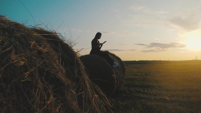 Girl farmer uses tablet in the field next to haystack at sunset. Smart farming, using modern technologies in agriculture.