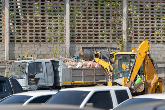 Yellow Digger Car Is Raising Up Polluted Waste Into Garbage Truck At Parking Lot With Cement Wall Behind.