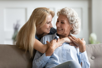 Smiling millennial girl hug cuddle mature mom relaxing together in living room, happy grownup adult daughter embrace senior 60s mother, rest enjoy family weekend at home, show love and care