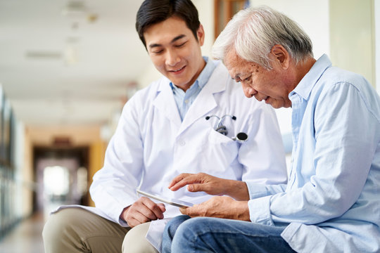 Young Asian Doctor Talking To Senior Man In Hospital Hallway