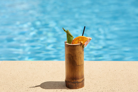 Outdoor picture of wooden cup with cocktail being near swimming pool, green and piece of orange being inside cocktail, black straw to drink, getting rid of thirst, cold drink in hot weather.