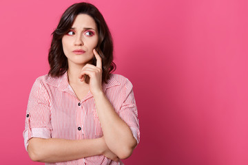 Indoor shot of attractive woman with thoughtful expression keeps hand under chin, looks aside, thinks about question, dressed striped blouse, posing isolated on pink background. Copy space for ad.