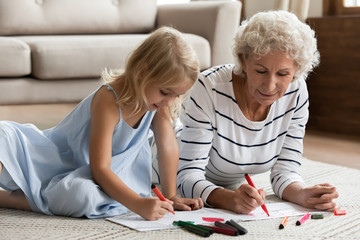 Happy loving mature grandmother lying on floor in living room drawing with cute little preschooler granddaughter, caring senior grandparent relax play have fun with small grandchild painting at home