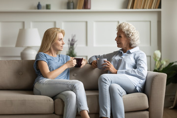 Happy senior mother and grownup daughter sit relax on couch in living room talk drinking tea or coffee together, mature mum and adult girl child rest on sofa, enjoy family weekend at home chatting