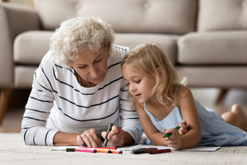 Caring senior grandmother lying on floor with cute little preschooler granddaughter drawing together, loving mature grandparent play have fun with small grandchild painting at home, education concept