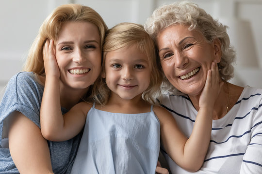 Portrait of cute little preschooler girl hug cuddle with happy young mother and mature grandmother at home, smiling three generations of women look at camera relaxing together, family bonding concept
