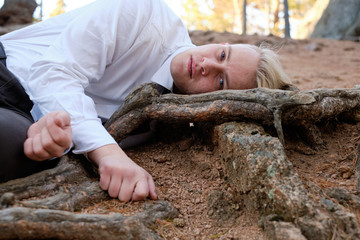 a man with blond hair in a white shirt and black coat is lying on the ground in the forest