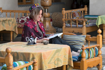 Young wooman in traditional uzbek coat (chapan) and scull cap with book by table with teapot and cup (piala). Uzbekistan, Central Asia.