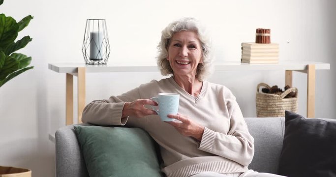 Smiling senior adult lady drinking tea looking away at camera at home. Cheerful retired old grey-haired woman laughing relaxing sitting alone on sofa in cozy living room. Happy elder grandma portrait