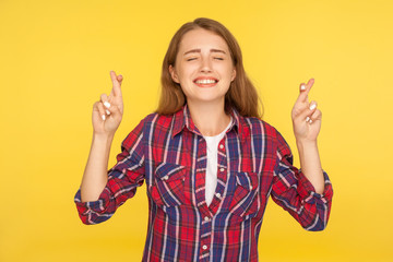 Portrait of cute hopeful ginger girl in shirt standing with crossed fingers and making wish,...