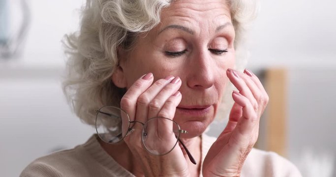 Tired senior adult woman taking off eyeglasses feeling eye strain concept. Exhausted older grandmother holding glasses massaging eyes having headache or bad vision, fatigue problem. Close up view