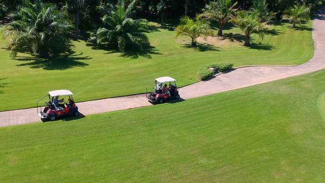 Two golf carts driving alongside tropical green golf course, aerial follow view
