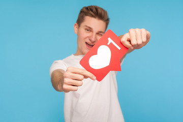 Cheerful smiling man in white t-shirt holding out social media button to camera, showing heart like icon, giving recommendation to support content, follow blog. indoor studio shot blue background