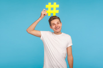 Media popularity. Carefree funny man in t-shirt holding big yellow hashtag symbol on his head and...