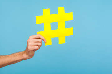 Closeup of male hand holding big yellow hash sign, hashtag symbol of internet trends and popular blogs, recommendation to follow social media content. indoor studio shot isolated on blue background