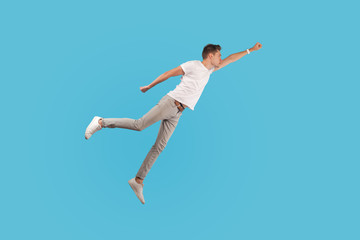 Full length portrait of motivated confident man in white t-shirt and casual pants flying up in air as superhero with raised hand, feeling superpower. indoor studio shot isolated on blue background