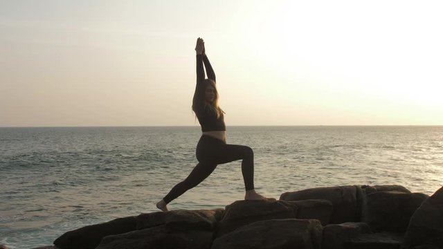 girl silhouette practices yoga exercises standing on large rocks against endless sea at sunrise slow motion. Concept body positive and healthy lifestyle