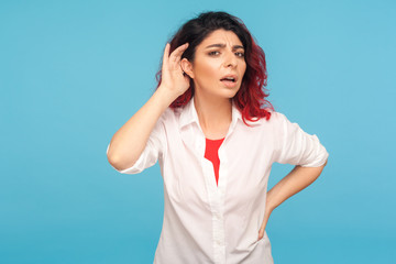 Portrait of nosy hipster woman with fancy red hair in shirt listening attentively holding hand near ear to hear confidential talk, hard to understand. indoor studio shot isolated on blue background