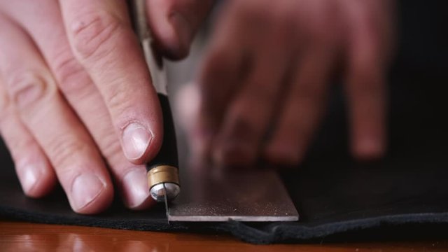 A tanner is cutting black leather sheet with a razor and metre. The master is making a leather belt for men.