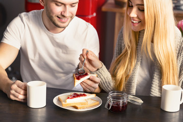 Happy guy and his girlfriend having toasts with jam and coffee for breakfast in kitchen