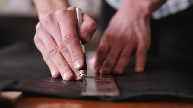 A tanner is cutting black leather sheet with a razor and metre. The master is making a leather belt for men.