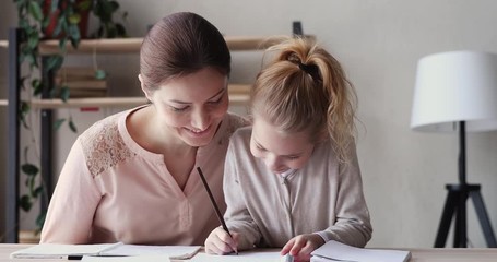 Happy young mum helping cute school child daughter doing homework together. Small 6-7 years kid girl learning writing in notebook studying with tutor. Children elementary handwriting education concept - Powered by Adobe