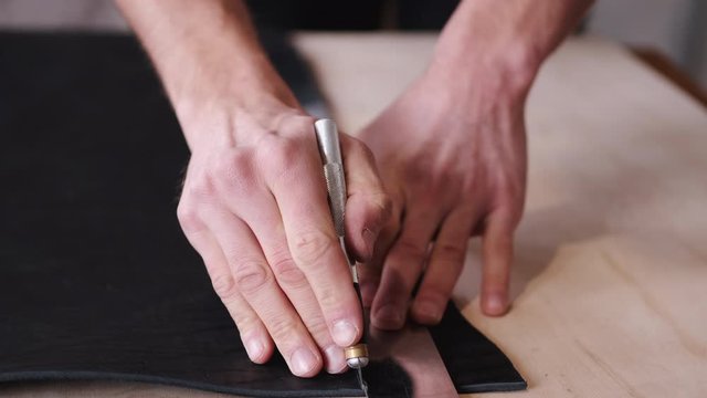 A tanner is cutting black leather sheet with a razor and metre. The master is making a leather belt for men.
