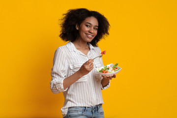 Smiling Afro Woman Eating Fresh Vegetable Salad Standing On Yellow Background © Prostock-studio