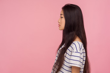 Side view of serious calm beautiful asian girl with long brunette hair in striped t-shirt looking at copy space with concentrated thoughtful expression. indoor studio shot isolated on pink background