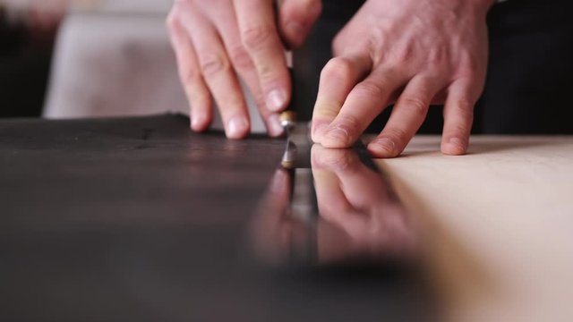 A tanner is cutting black leather sheet with a razor and metre. The master is making a leather belt for men.